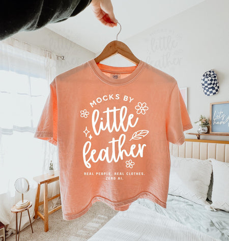 a peachy comfort colors 3023cl t-shirt with bold white text that says 'Mocks by Little Feather – Real People. Real Clothes. Zero AI.' surrounded by doodle-style flowers and feathers. Trendy Etsy-style mockup featuring real model photography for print-on-demand sellers.