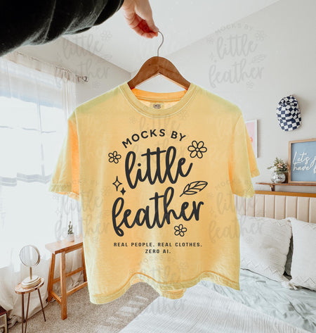 a butter comfort colors 3023cl t-shirt with bold white text that says 'Mocks by Little Feather – Real People. Real Clothes. Zero AI.' surrounded by doodle-style flowers and feathers. Trendy Etsy-style mockup featuring real product photography for print-on-demand sellers.