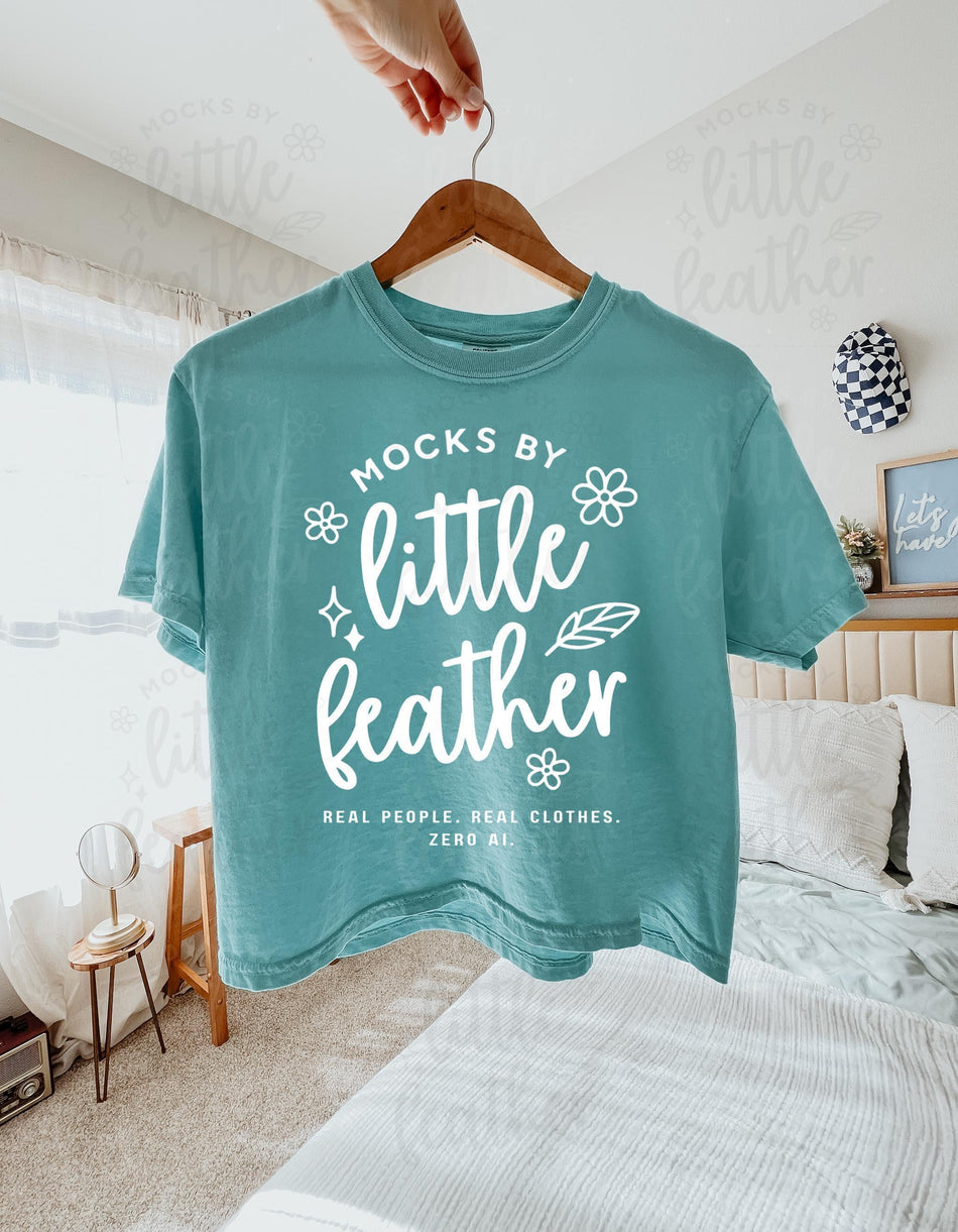 a chalky mint comfort colors 3023cl t-shirt with bold white text that says 'Mocks by Little Feather – Real People. Real Clothes. Zero AI.' surrounded by doodle-style flowers and feathers. Trendy Etsy-style mockup featuring real model photography for print-on-demand sellers.