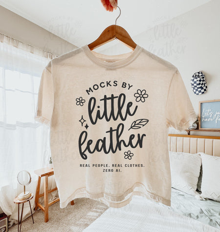 An ivory comfort colors 3023cl t-shirt with bold white text that says 'Mocks by Little Feather – Real People. Real Clothes. Zero AI.' surrounded by doodle-style flowers and feathers. Trendy Etsy-style mockup featuring real product photography for print-on-demand sellers.