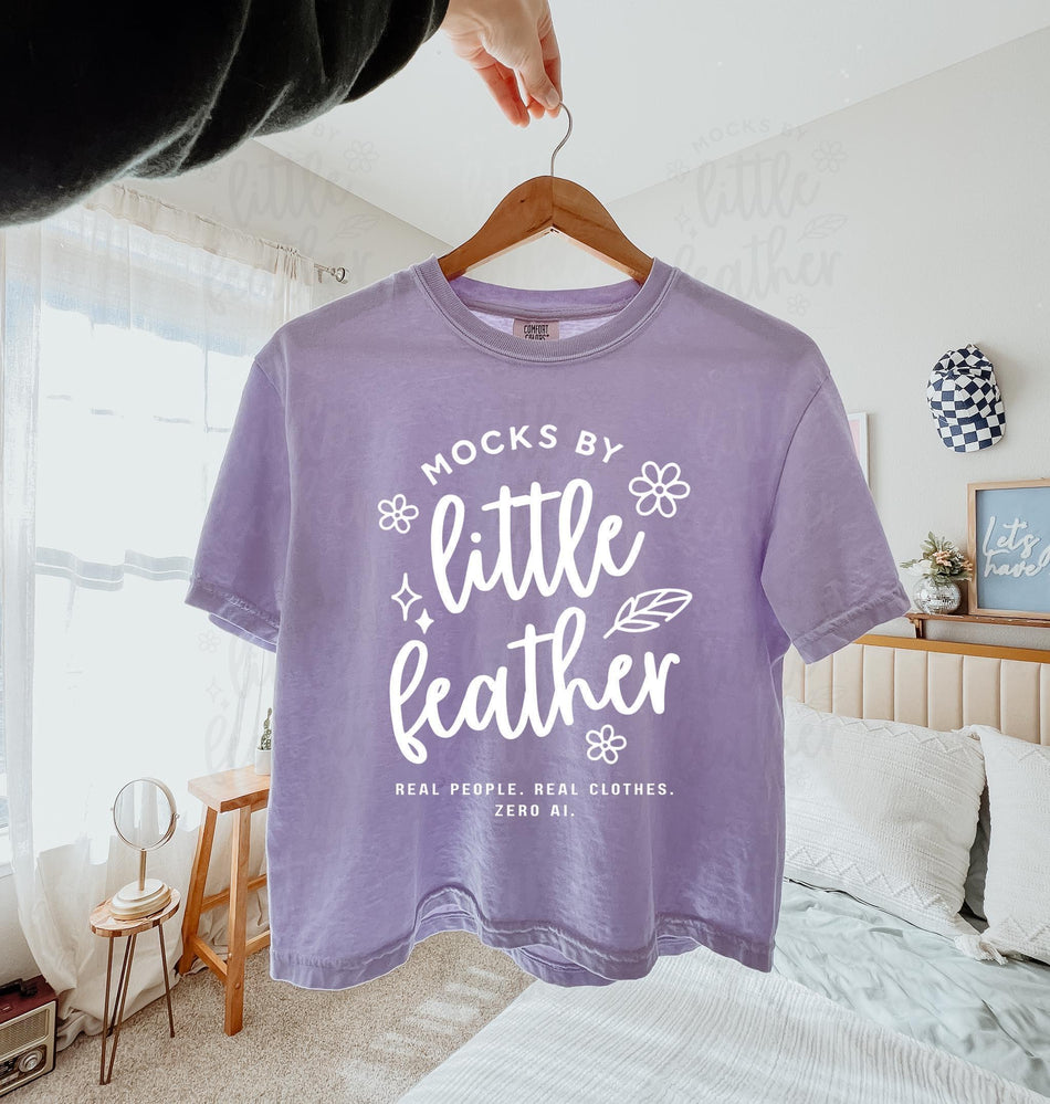 orchid comfort colors 3023cl t-shirt with bold white text that says 'Mocks by Little Feather – Real People. Real Clothes. Zero AI.' surrounded by doodle-style flowers and feathers. Trendy Etsy-style mockup featuring real product photography for print-on-demand sellers.
